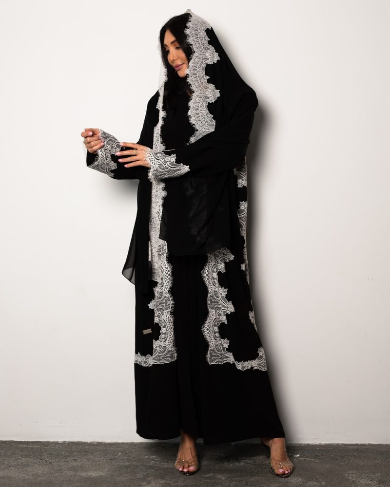 Black abaya with white lace trim, full-length shot against a white wall